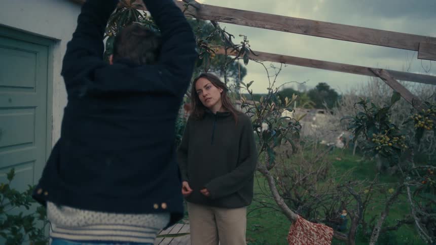 A woman and a man are in a backyard surrounded by various plants and the woman is looking at her nails