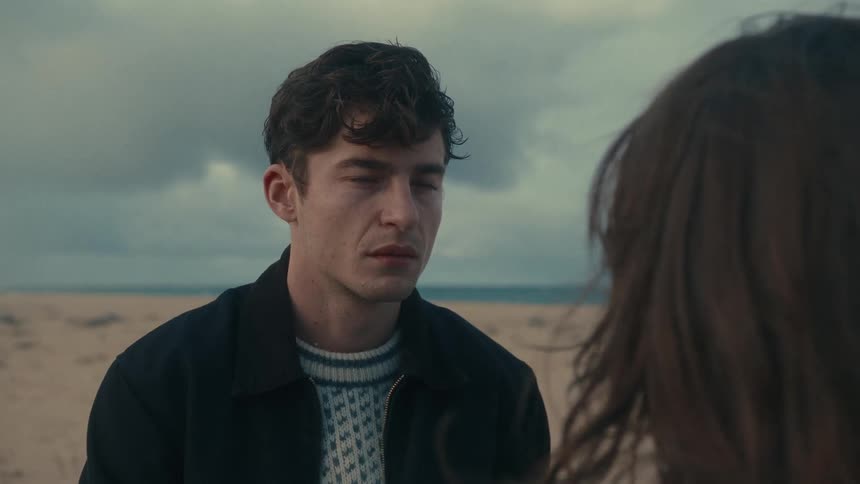 Man with a serious expression talks to a woman on the beach under a cloudy sky