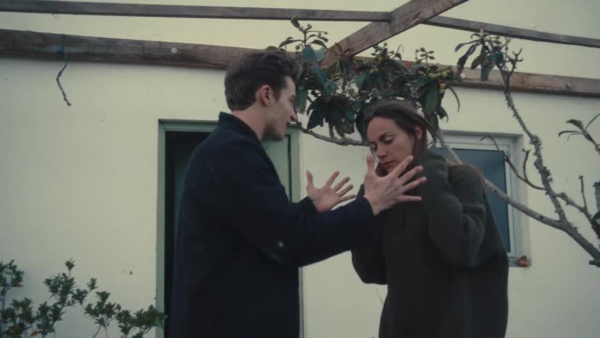 A male and female stand outdoors near a white house in an argument the male is angry and the female is nervous
