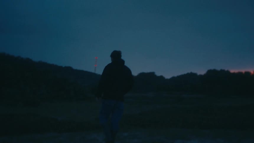 Seen from behind a mans silhouette walks slowly across a dark field as the last light fades on the horizon against a deep blue twilight sky