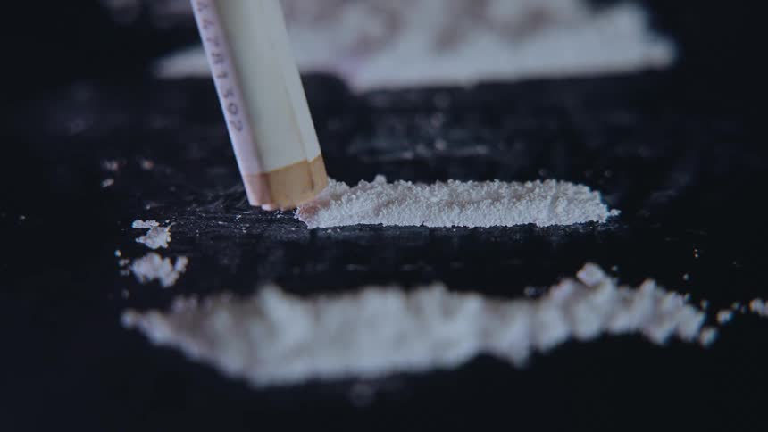 A pile of white powder is next to 2 lines of powder one line is being sucked up with a paper straw on a black surface