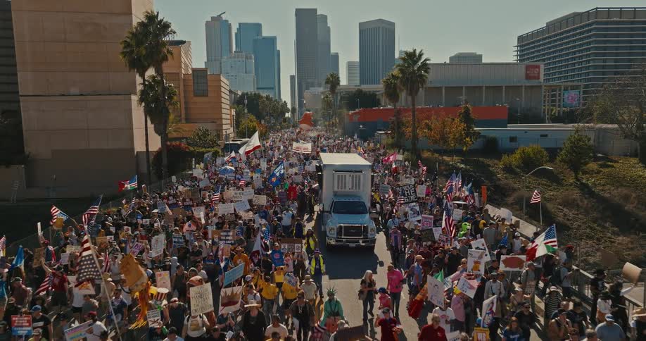 Crowd marches down a wide avenue holding signs and flags with skyscrapers in the background