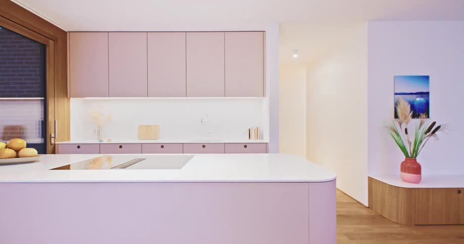A slow pan reveals a sleek modern kitchen with pastel pink cabinets and a matching island moving past a fruit bowl to show a bright adjoining hallway