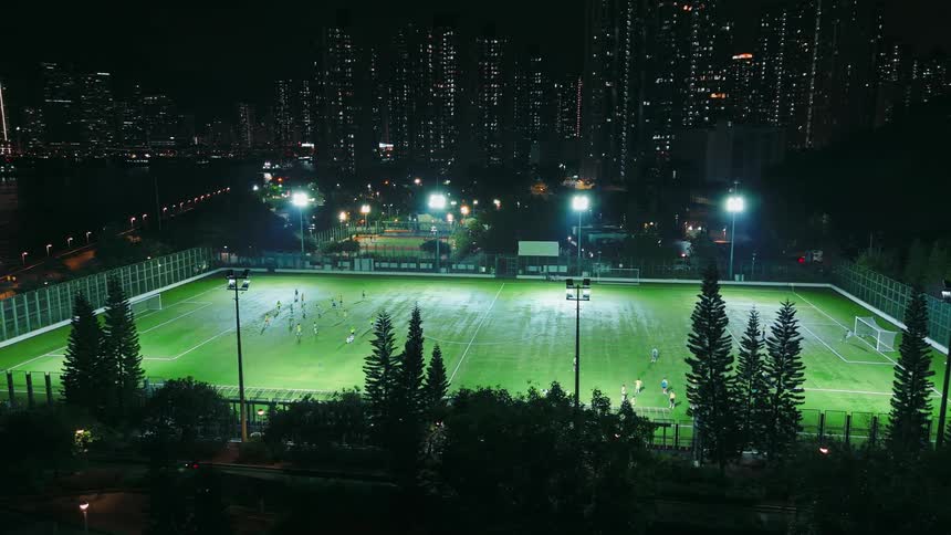 People play a soccer match at night on a brilliantly illuminated field set against the backdrop of a dense glittering city skyline