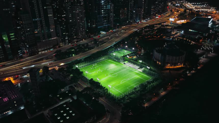 Traffic traveling on a divided freeway passes between the city skyscrapers and a waterfront soccer field at night