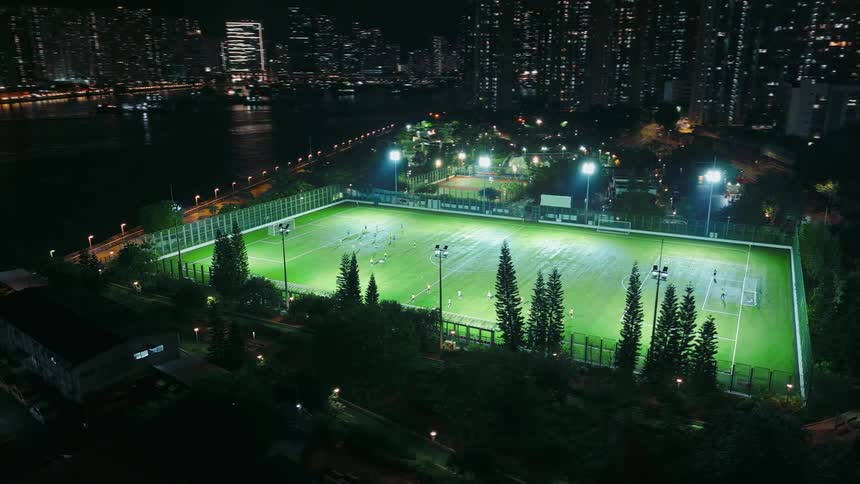 A brightly illuminated artificialturf soccer pitch where several individuals are actively playing situated in a small urban park surrounded by dark dense trees with a waterfront and a backdrop of towering residential and commercial buildings