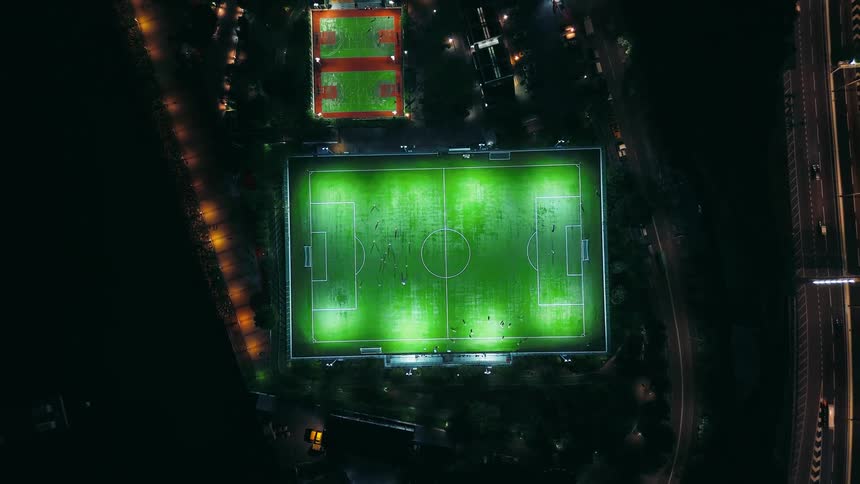 A brilliantly illuminated green turf soccer field at night where players are actively moving standing out sharply against the encompassing darkness of the surrounding park and the neighboring streaked lights of a multilane highway