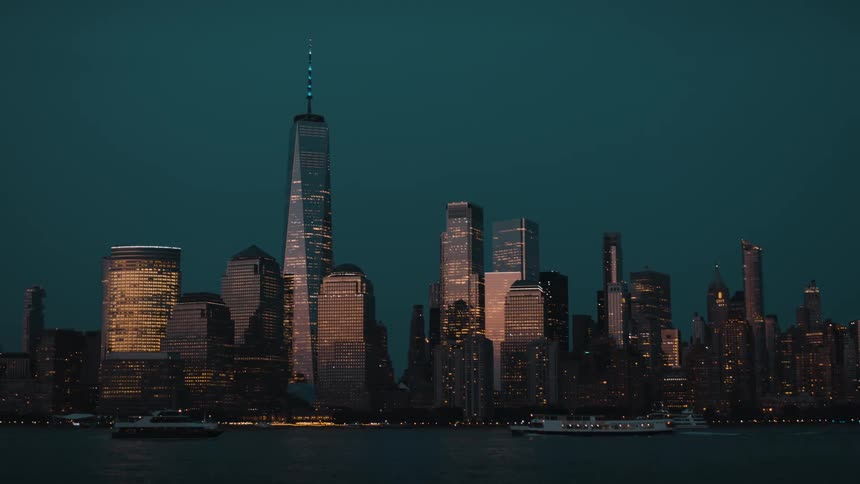 The video presents a striking view of a large city skyline dominated by tall illuminated skyscrapers against a dark twilight sky in the foreground the water reflects the lights of the buildings and a few boats can be seen moving across the surface