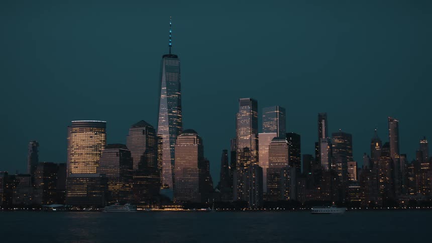 The towering skyscrapers of the lower manhattan skyline glow with thousands of lights against the deep blue twilight sky as boats pass by on the water below
