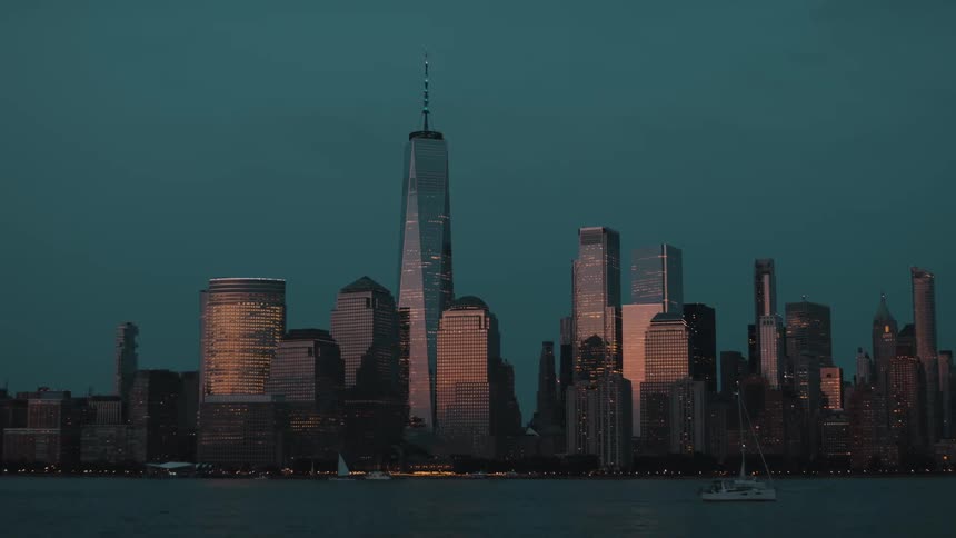 A wide shot with several building silhouettes against a dark teal twilight sky with many buildings glowing from within all viewed from across a body of water where small sailboats floats in the foreground