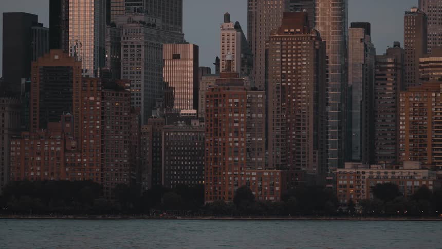 A slow pan moves across a dense city skyline bordering a dark river capturing countless skyscrapers and buildings standing against the hazy dusk sky as lights begin to glow