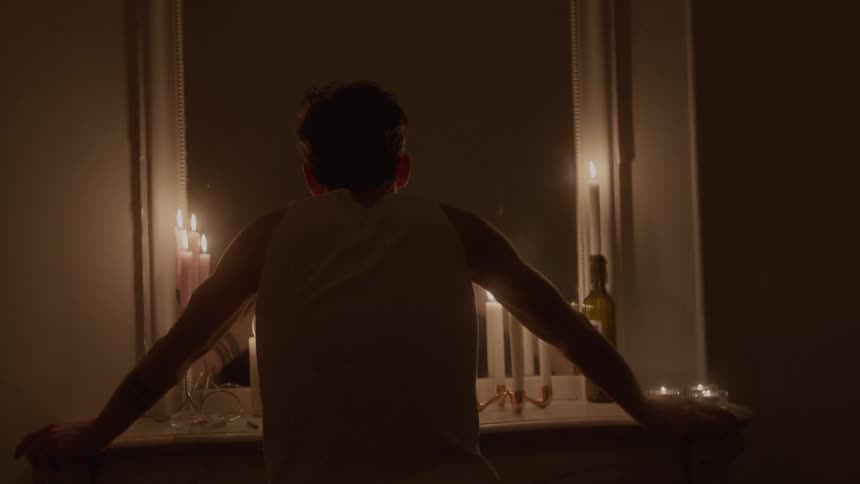 The video shows a man standing with his back to the camera in a dimly lit room leaning on a mantelpiece or shelf wearing a lightcolored tank top or undershirtthe mantelpiece is decorated with numerous lit candles and a tall mirror behind him
