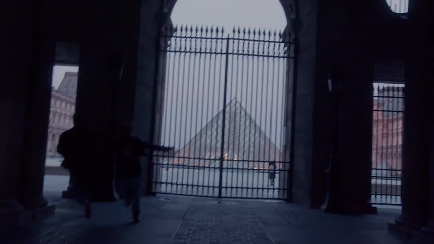 From a dark stone passageway two figures run past a large iron gate that opens onto a view of the louvre pyramid on an overcast day