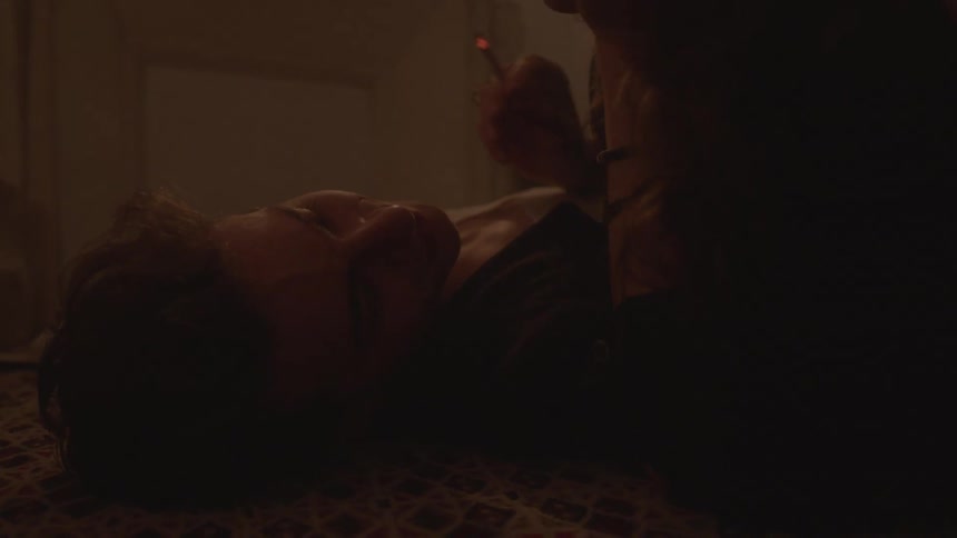 A young couple lies on the floor of a dimly lit room illuminated by candlelight from a nearby shelf as one of them smokes a cigarette
