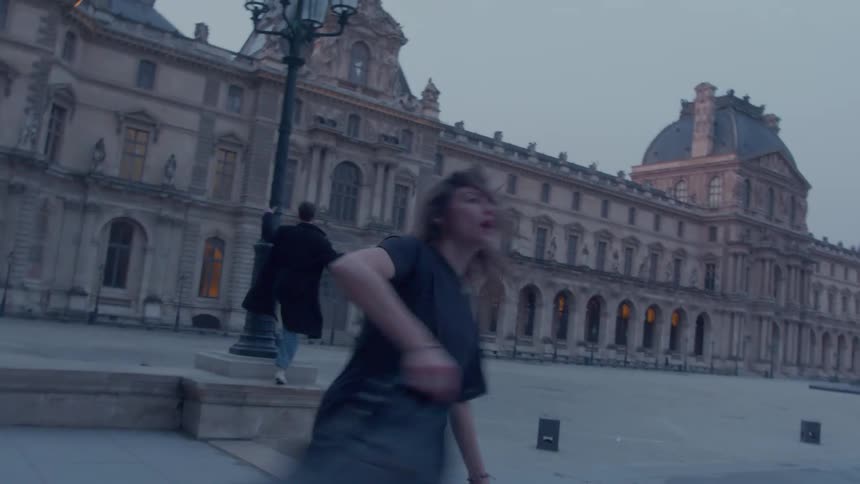 Woman runs smiling with a jacket in hand in front of a historic building at dusk
