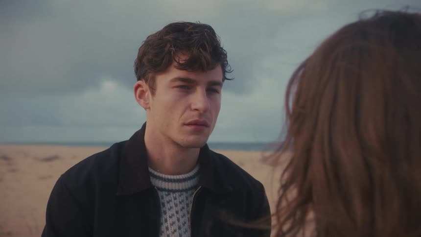 Man faces a woman on the beach with a serious expression under a cloudy sky