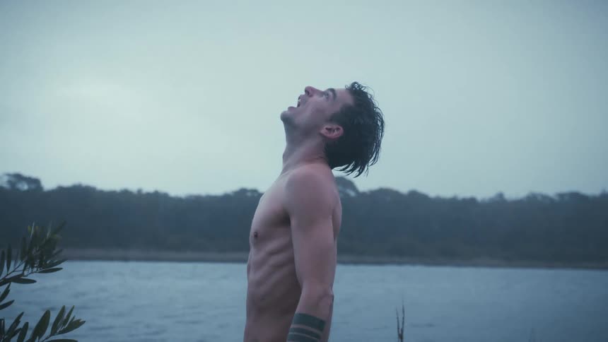 Shirtless man screams looking upward by the edge of a gray lake