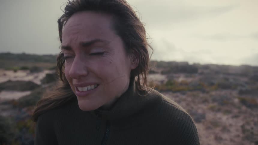 In a desolate landscape a woman with windblown hair transitions from a pained smile to a look of profound sorrow as the camera pushes in on her face