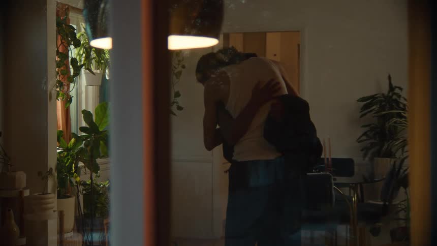 A woman enters a living room and a man who is on the sofa gets up and hugs her
