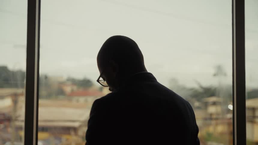 A man stands at a large window while talking on his phone