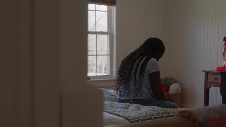 A young person sits sadly on a bed in a sunlit bedroom