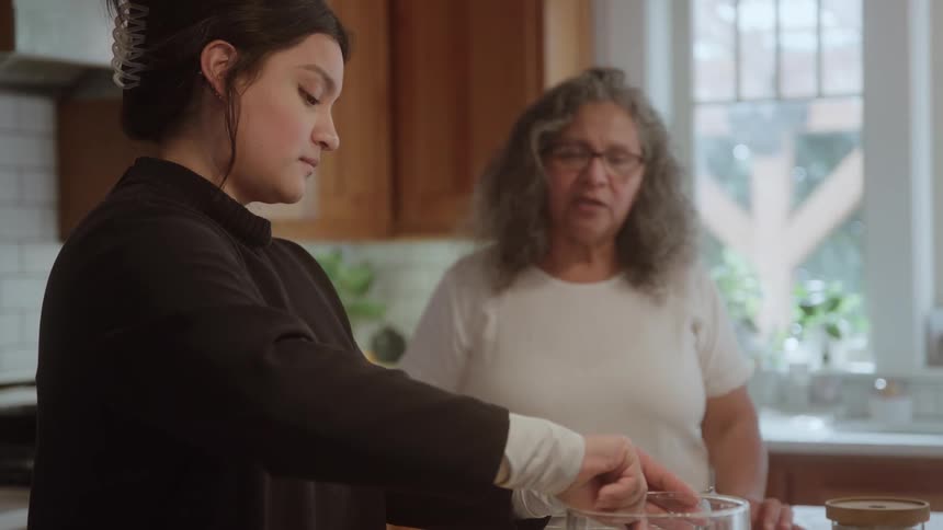 A young woman pours an ingredient from a measuring cup into a glass bowl as an older woman watches and talks with her baking together in a bright sunlit kitchen