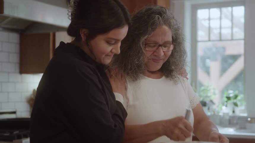 Elderly woman mixing ingredients in a bowl while a young woman hugs her while talking and smiling indoors during day time
