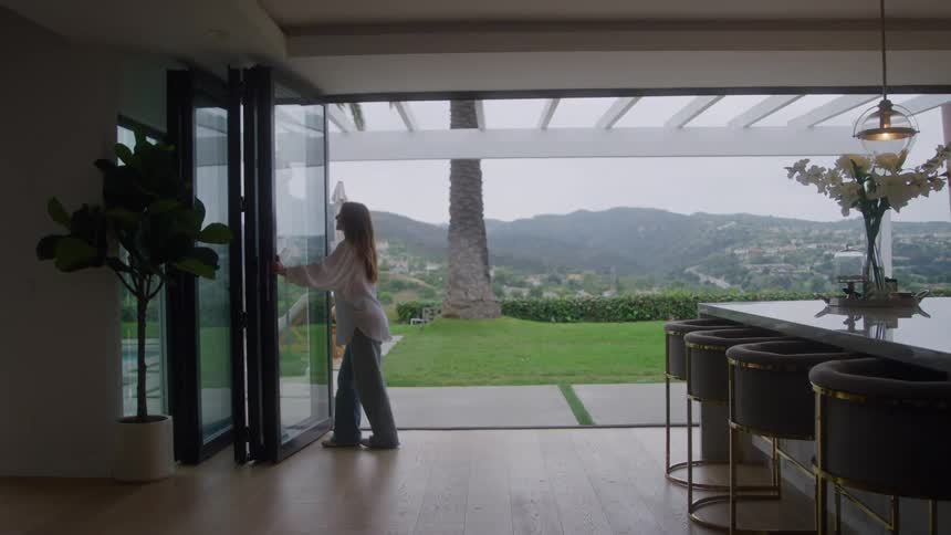 A woman wearing a white shirt and blue pants slides open a large folding glass door in a modern kitchen with bar stools revealing a green lawn and a view of rolling hills under an overcast sky