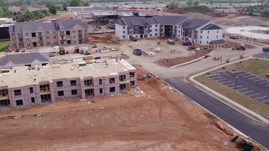 The video shows an aerial view of an active construction site for a large residential or apartment complex with buildings in various stages of completionthe foreground features a building structure framed but not yet fully finished surrounded by bare