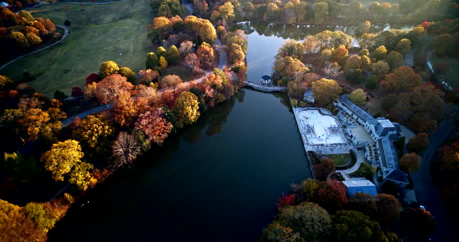 During autumn a calm reflective lake is surrounded by colorful trees in shades of orange yellow and redthere is a white boathouse with a dock and a grassy area with buildings are on the left