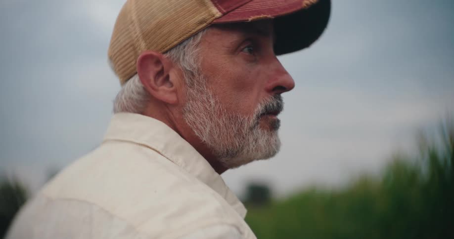 A thoughtful grey bearded farmer in a cap looks off into the distance