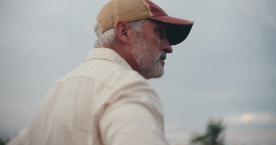 A grey bearded farmer in a cap looks up at the sky with a contemplative expression while standing in a field