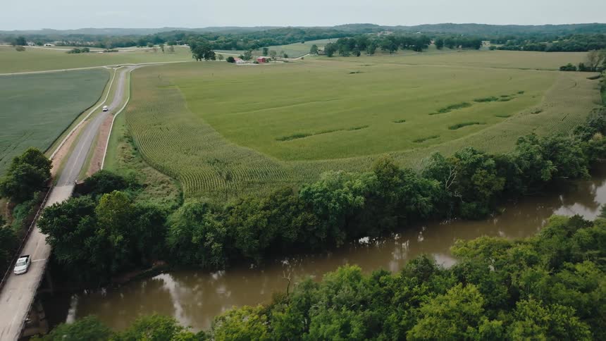 An aerial view of a small bridge over a muddy river beside green farmland
