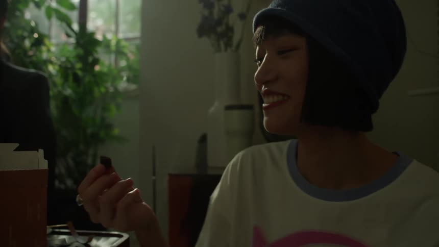 Three friends laugh and share snacks in a sunlit room with one playfully tossing a piece of food into her mouth