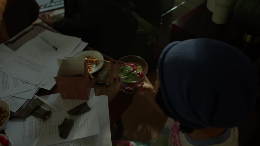 An overhead view shows one person in a blue hat holding a cocktail and another woman smiling as the group of friends sits on the floor around a low table piled with papers snacks and a recording microphone