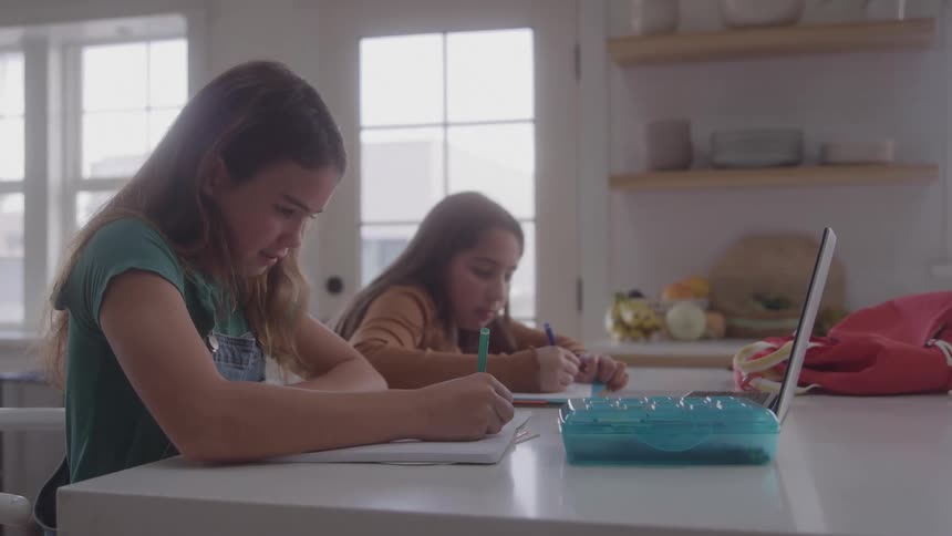 Two girls write in their notebooks while sitting at the kitchen table