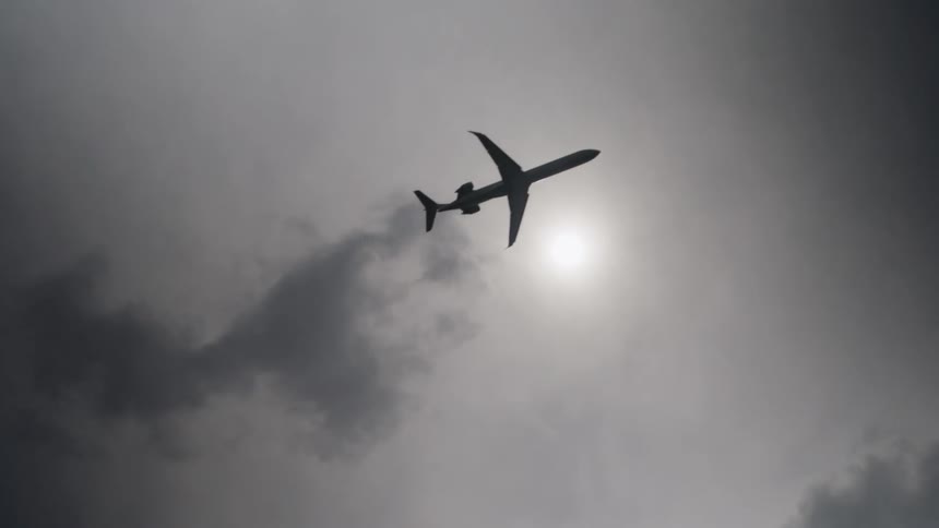 A dark silhouette of an airplane flies diagonally across a grey overcast sky passing through hazy clouds and past the bright diffused sun