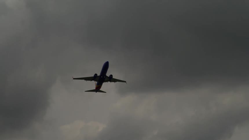 A passenger jet climbs steeply into a heavy overcast grey sky seen from below as it flies directly over a street intersection with red traffic lights