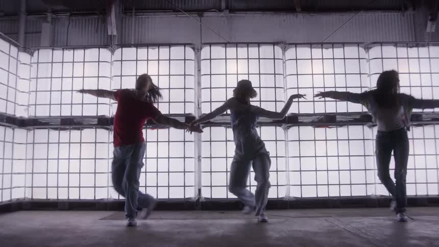 Three dancers perform a synchronized hip hop routine in an industrial warehouse silhouetted against a wall of bright glowing cubes