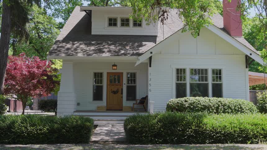 The facade of a wellkept twostory white house with a dark roof and a small covered porch featuring a wooden front door all framed by mature green trees and manicured shrubbery under bright sunlight