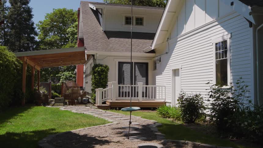 The camera moves slowly forward along a stone path in a sunny backyard gliding past a simple disc tree swing towards the back porch of a white house