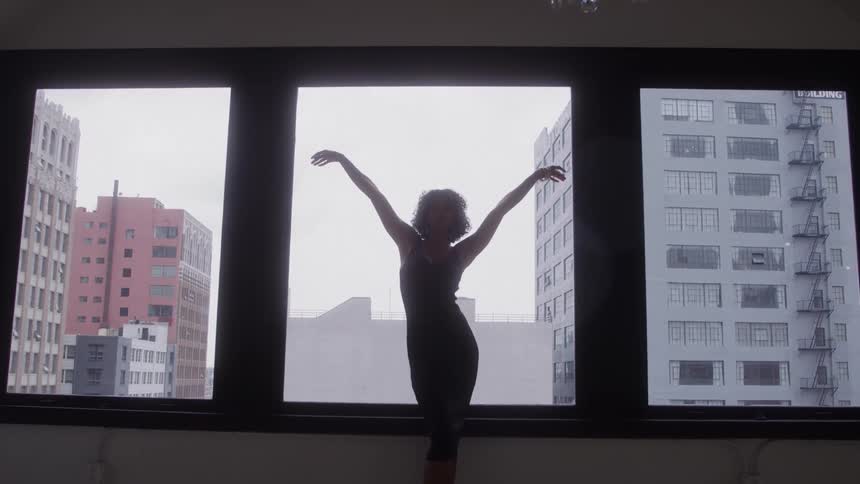 Woman standing partially silhouetted against a window in a downtown building raises her arms above her head while posing