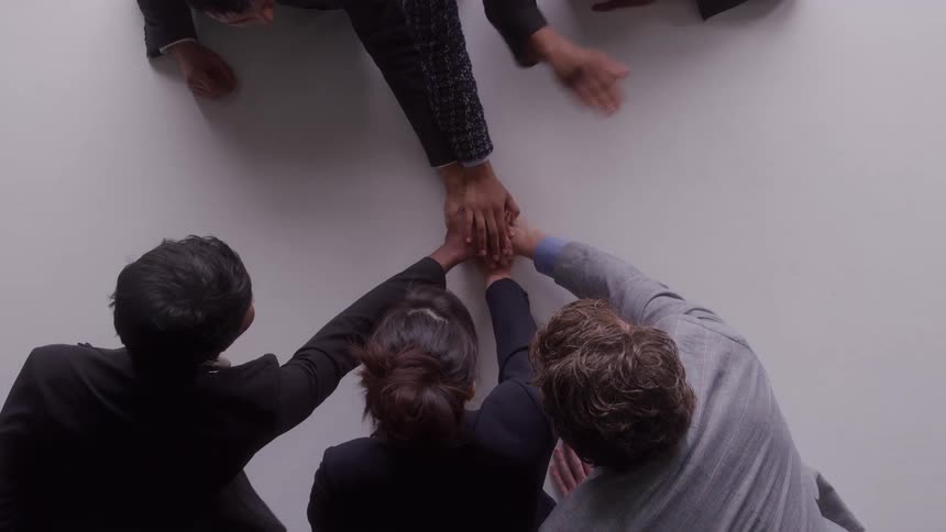 Five people reach their hands toward the center of a white table stack them in a collaborative team gesture and then quickly pull them back apart