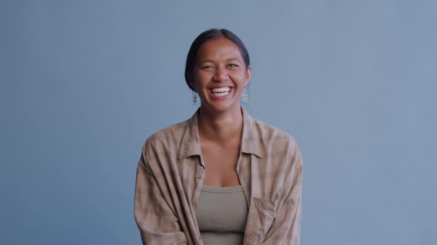 A young woman with dark hair pulled back wearing a neutraltoned plaid shirt over a tank top positioned against a plain bluegray backdrop as she bursts into wide genuine laughter with her eyes closed and teeth showing