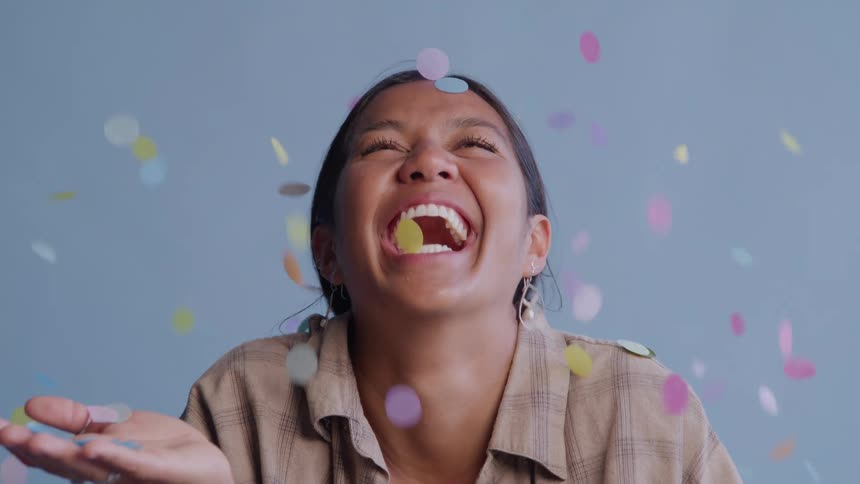 A young female from the chest up is laughing as confetti rains down on her and sticks to her mouth against a blue background