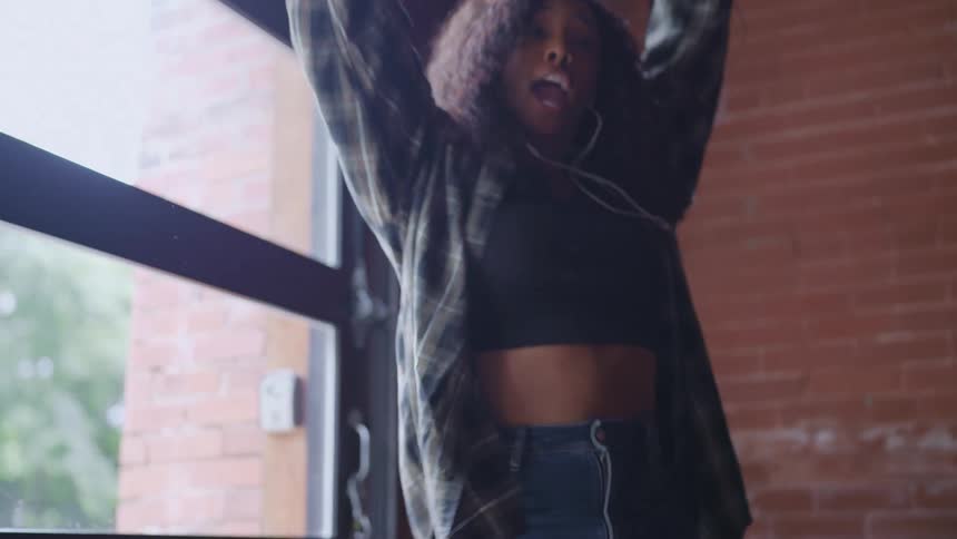 A young woman with long dark curly hair wearing a crop top an unbuttoned plaid shirt and jeans dancing with her arms raised all while wearing wired earbuds illuminated by bright natural light streaming in from a large industrialstyle window