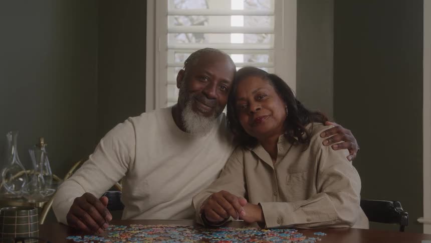 A black female and male sit at a dining room table hugging and sitting close appearing romantic