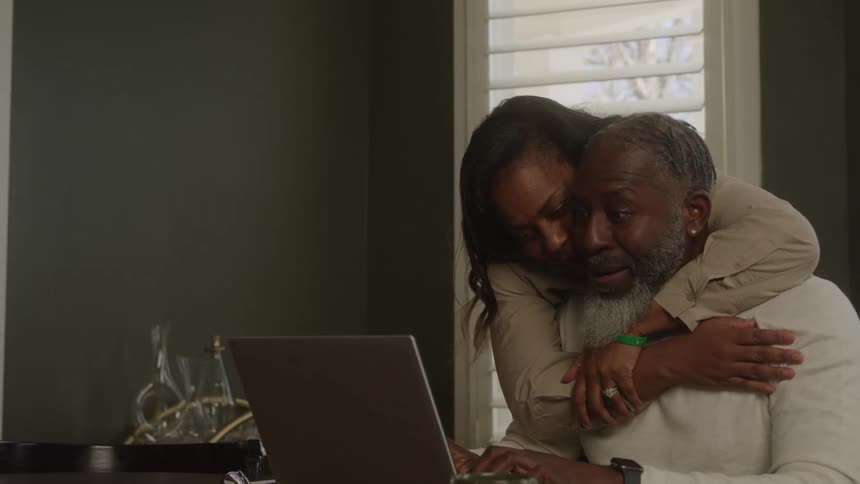 A wife hugs her husband while he sits and types on his laptop