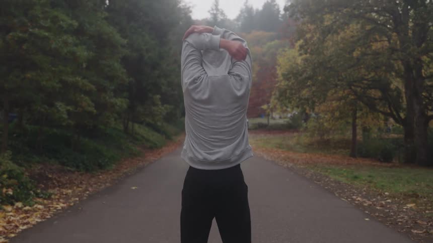 A man in a grey hoodie stretches his arms while standing in the middle of a paved path in a park on a cool day