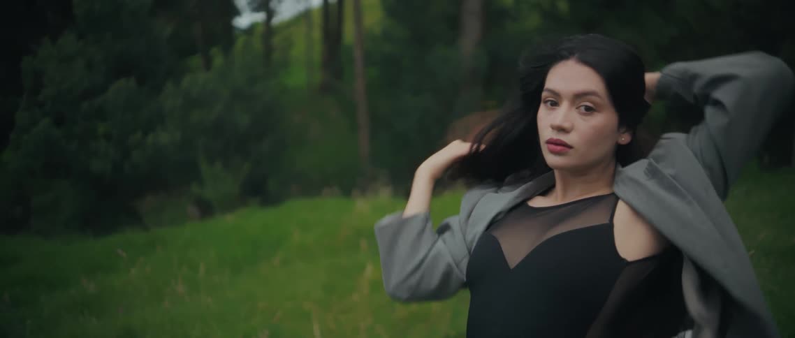 A young woman with long dark hair wearing a grey jacket moves her arms around her head while looking thoughtfully forward in a lush green outdoor setting
